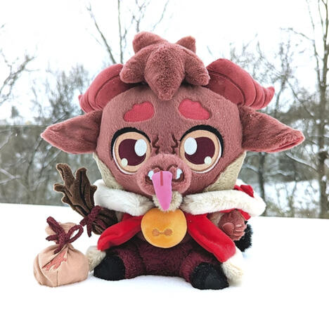 Kidd Krampus Plushpanion: A brown goat-like plush sits in the snow. Wearing his lush red winter cloak (hood down) and yellow jingle bell necklace. He hold his magnet twig prop in his hand and his magnetic tongue at his mouth. Kidd has yellow eyes, dark red