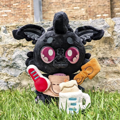 Monty Mothman Plushpanion: A soft black mothy plush sits in the grass. He is wearing his tan artist's apron and holding a thermometer and wooden spatula plush prop. In front of him sits his other two magnetic plush props, a candle and measuring cup.